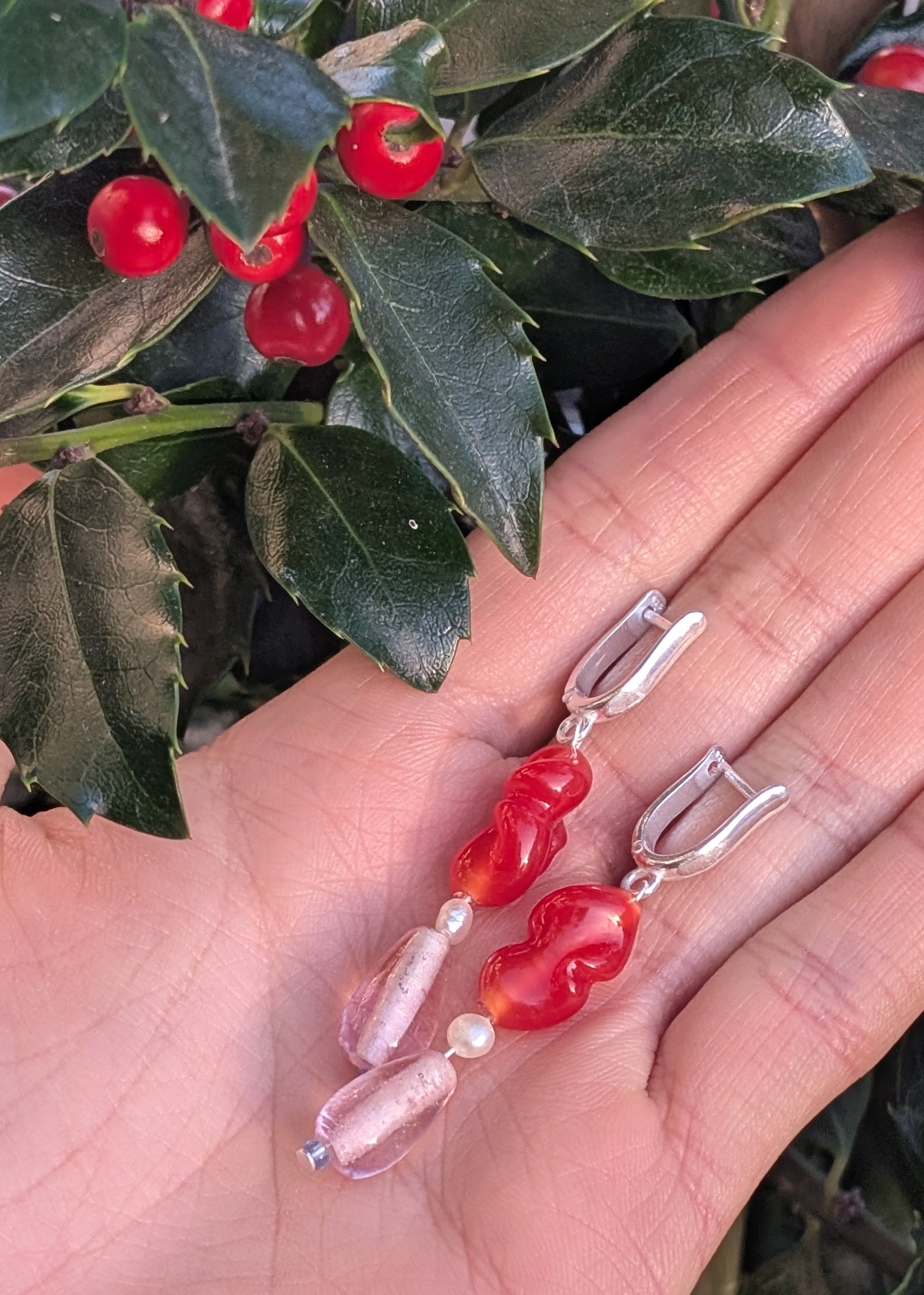Hand holding two earrings with red and pink beads against a green leafy background