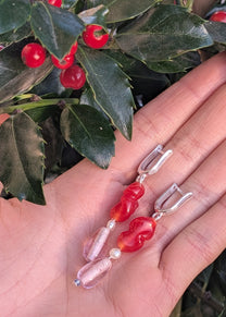 Hand holding two earrings with red and pink beads against a green leafy background