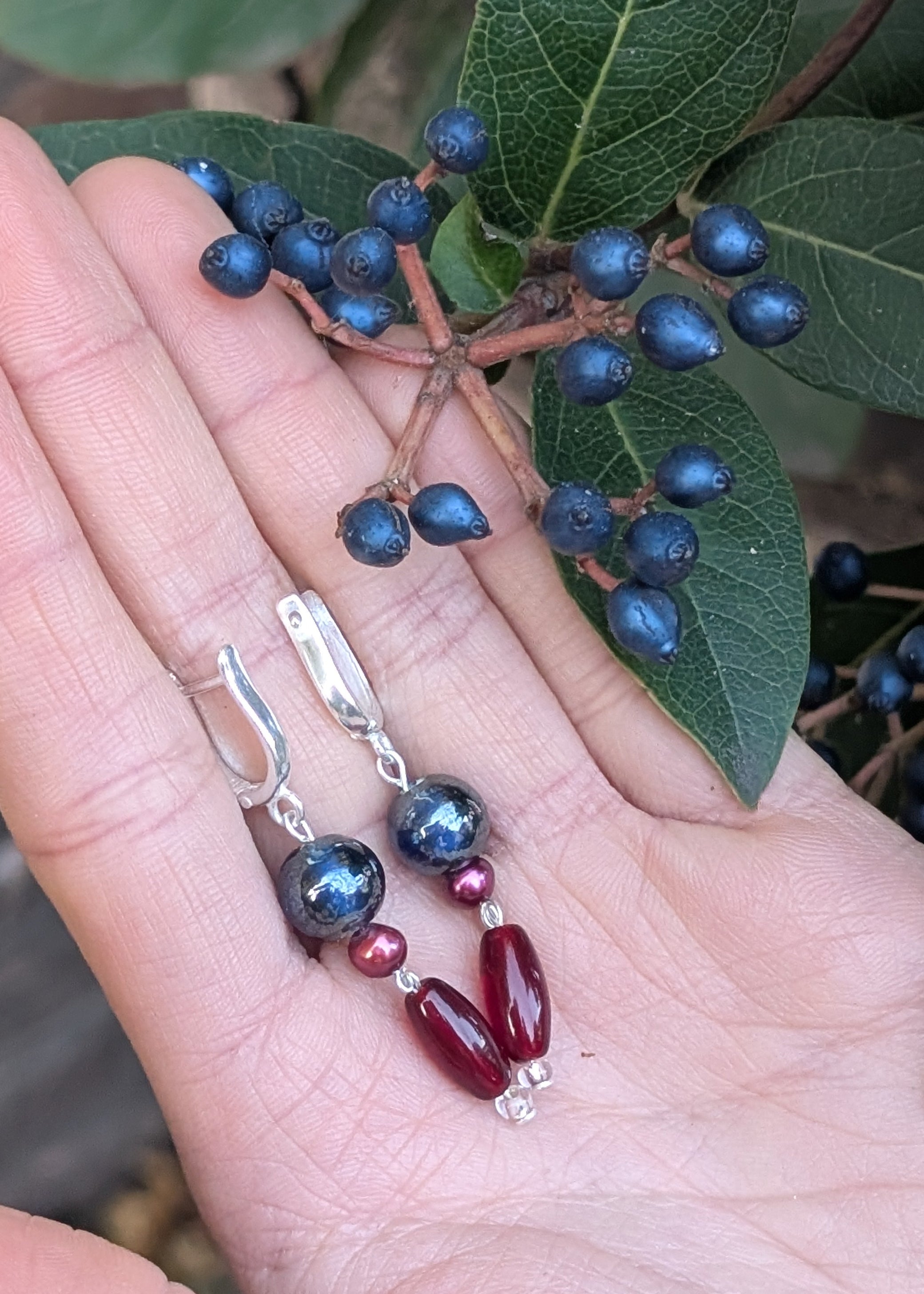 Silver earrings with blue and red beads held in a hand with green leaves in the background