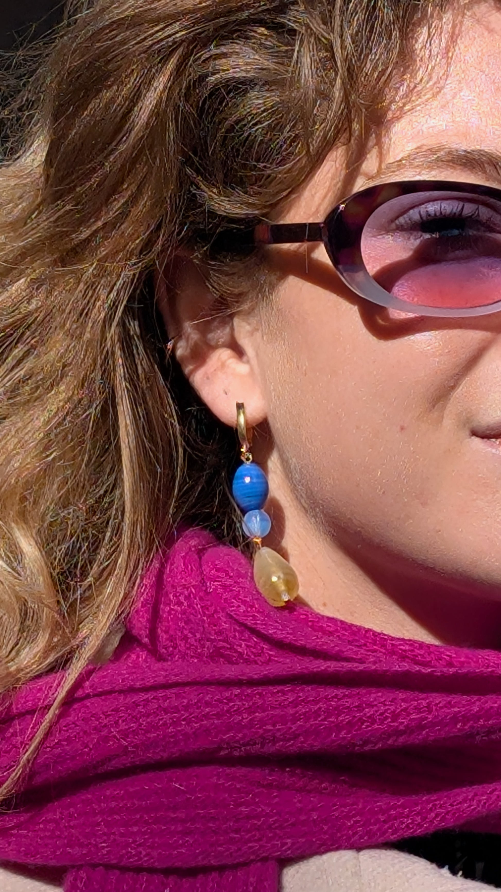 Close-up of a person wearing colorful earrings and sunglasses with a pink background.