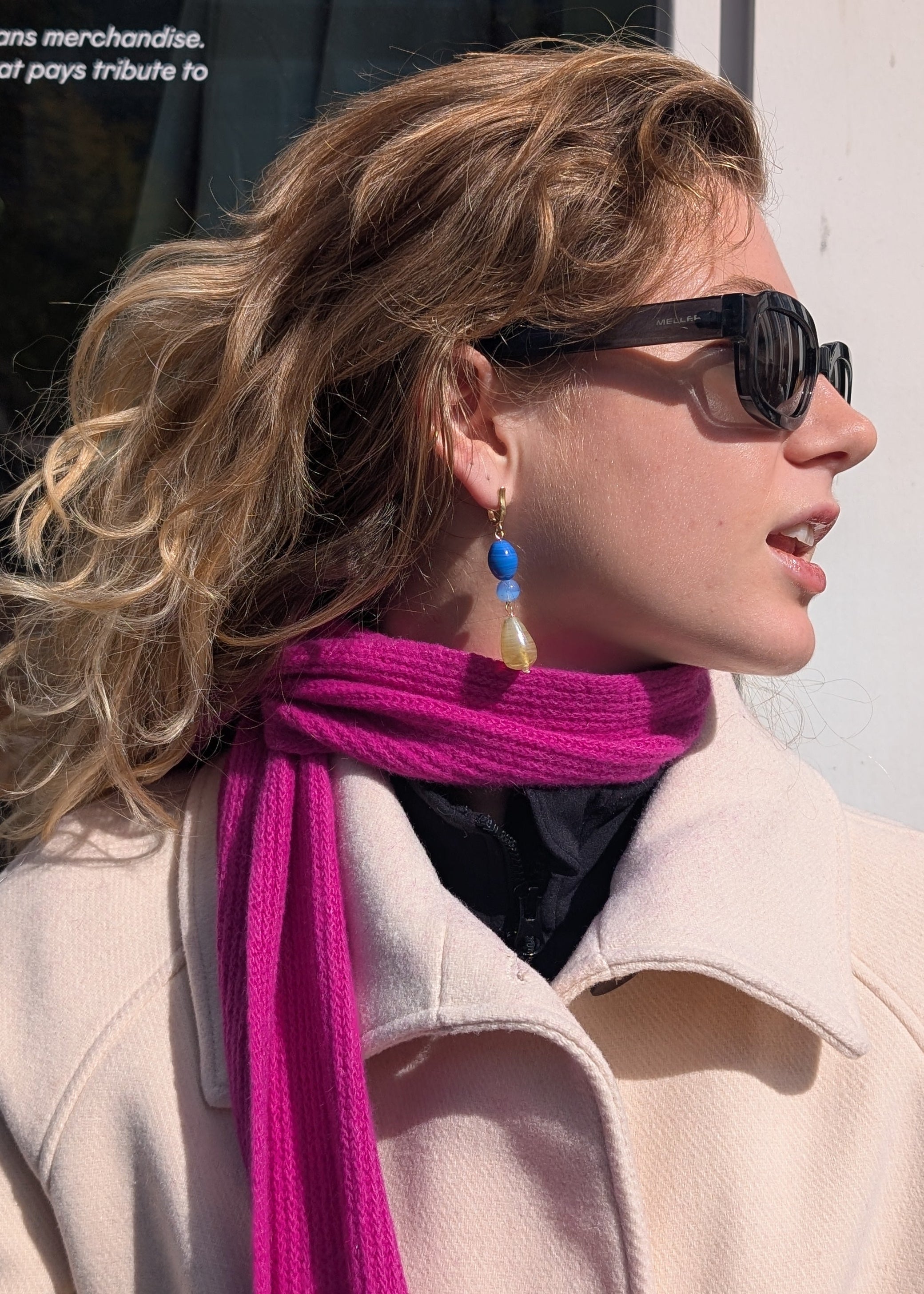 Woman wearing sunglasses and a bright pink scarf with a beige coat.