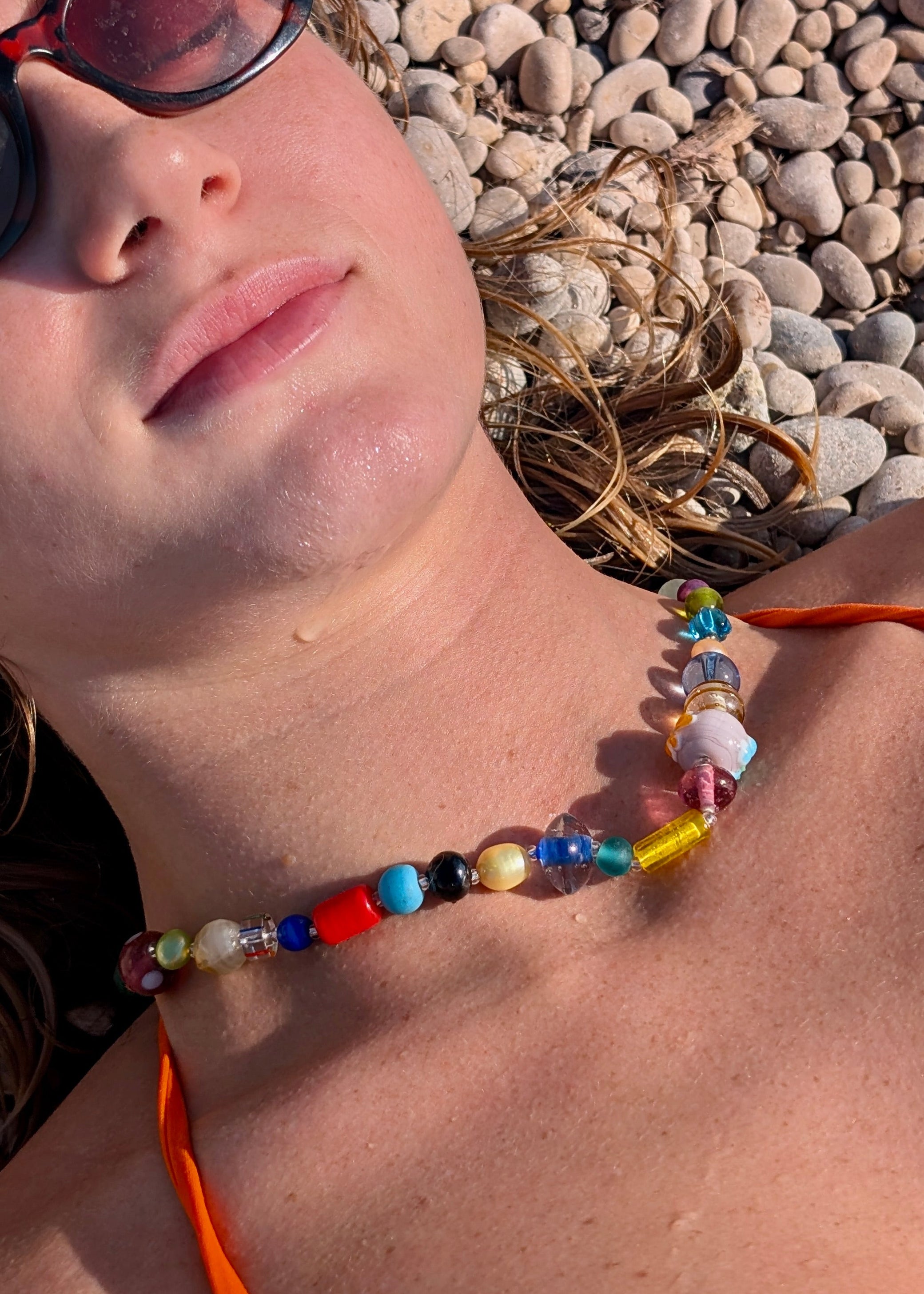 Woman wearing sunglasses and a colorful beaded necklace lying on pebbles.