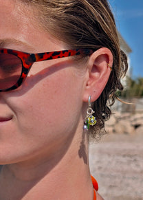 Close-up of a person wearing tortoiseshell sunglasses and colorful earrings with a blurred outdoor background.